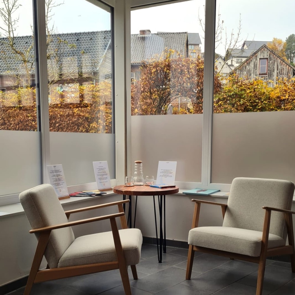 Zwei Sessel und ein kleiner Tisch am Fenster der Praxis in Nettersheim, dahinter herbstliche Bäume
