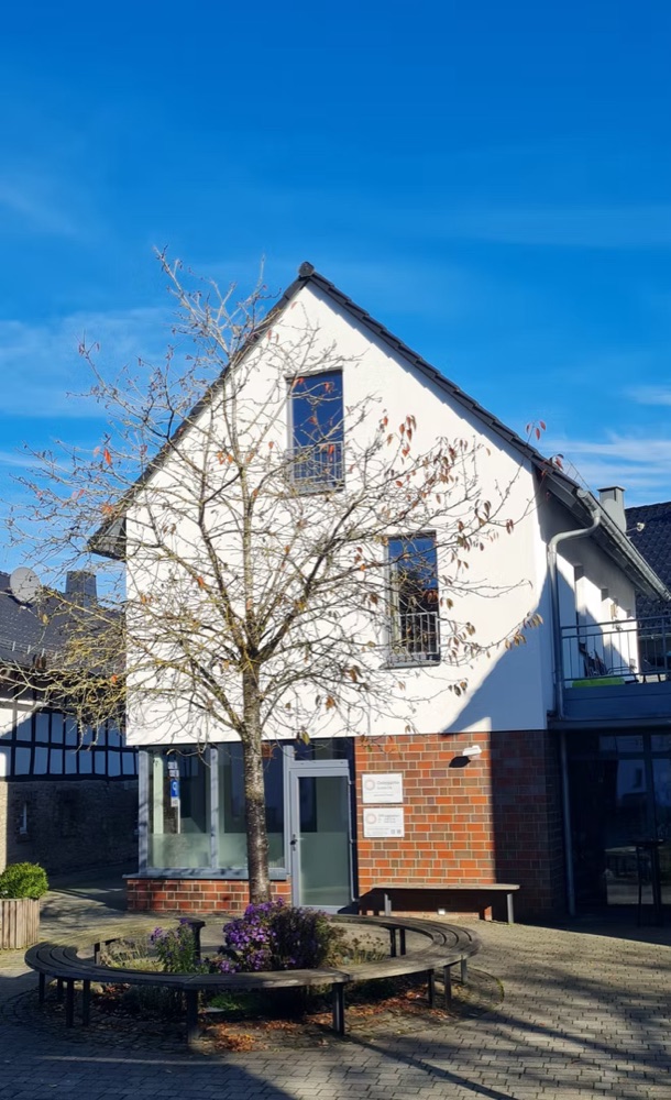 Praxisgebäude in Nettersheim, weißes Giebelhaus mit Backsteinanbau und Bank davor